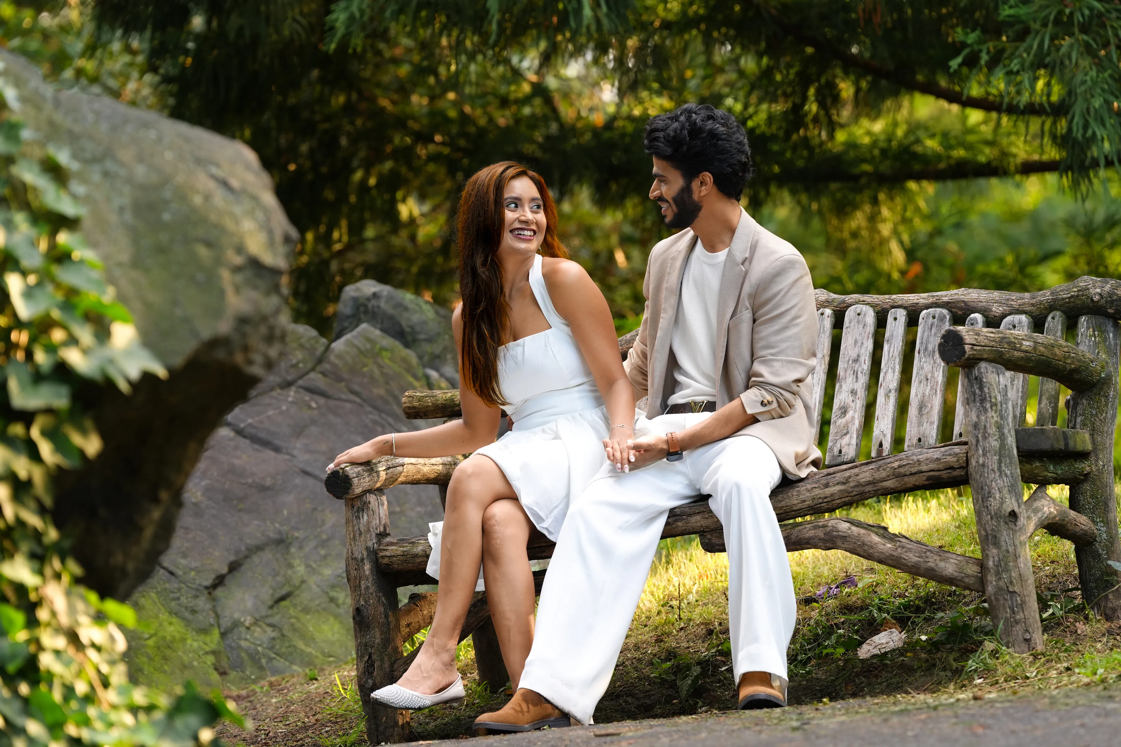 A couple seated together on a rustic park bench, laughing, framed by summer greenery